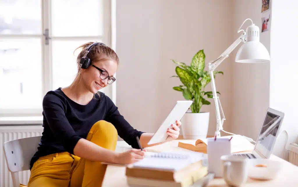 A young happy college female student sitting at the table at home, using tablet and headphones when studying.